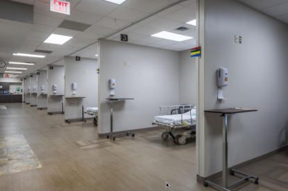 A hospital hallway with new flooring installation, empty patient bays, beds, and wall-mounted sanitizer dispensers under fluorescent lighting.