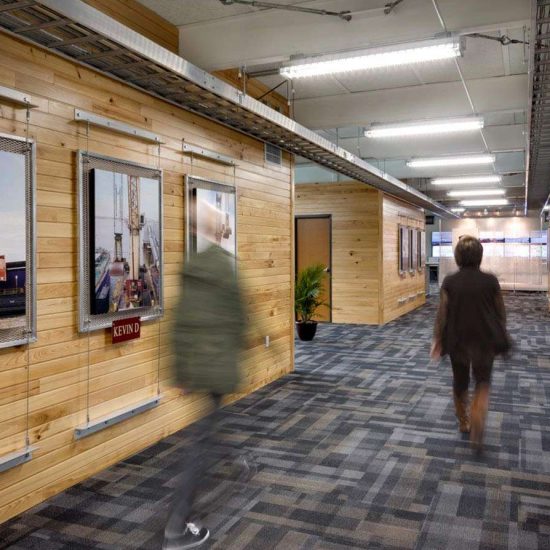 Two people walk down a hallway with wood-paneled walls, framed industrial photos, and new flooring installation underfoot.