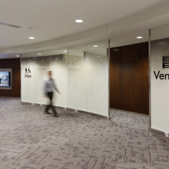 A person walks past men's restroom signs and vending machines in a modern hallway with carpeted floors by a flooring installation company.
