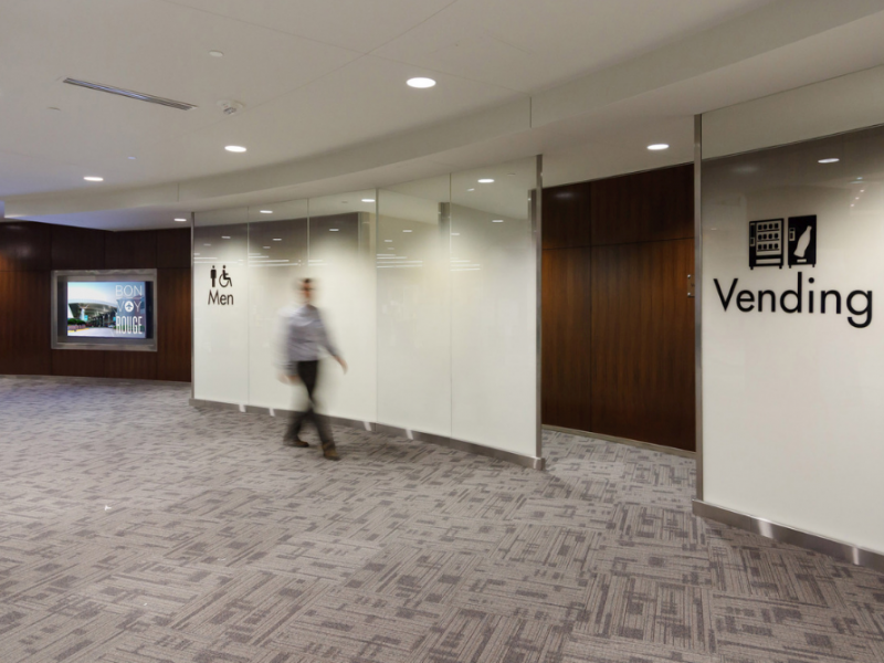 A person walks past men's restroom signs and vending machines in a modern hallway with carpeted floors by a flooring installation company.