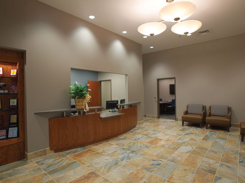 Reception area with a wooden desk, awards, chairs, and tiled floor by a flooring contractor in a modern office setting.