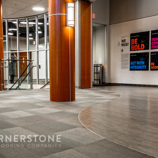 Lobby with gray carpet and tile, wood columns, glass railing, and motivational posters. “Cornerstone Flooring Companies,” a commercial flooring company, logo visible.