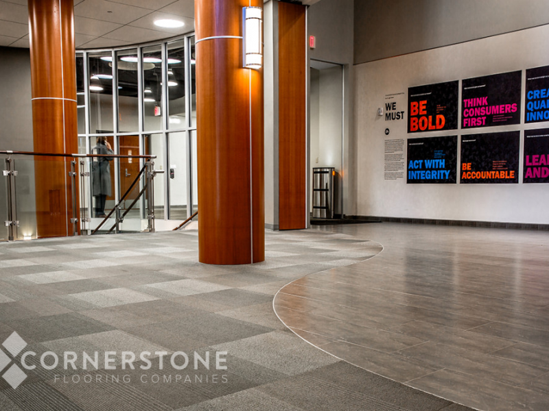 Lobby with gray carpet and tile, wood columns, glass railing, and motivational posters. “Cornerstone Flooring Companies,” a commercial flooring company, logo visible.