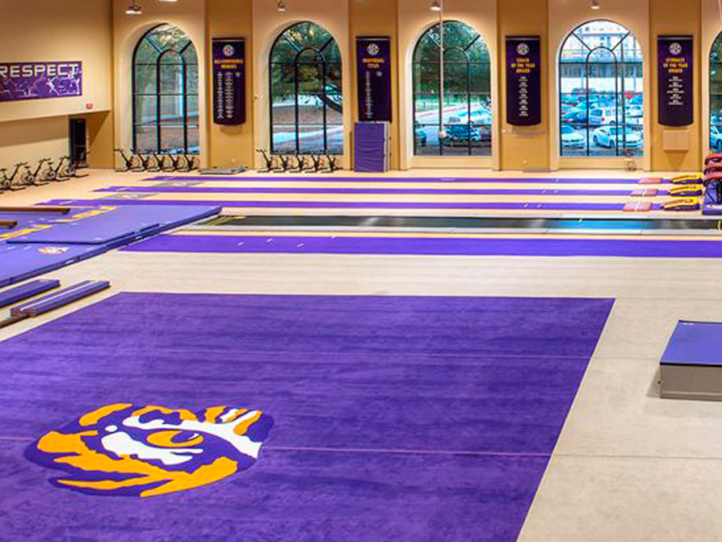 Large indoor gym with new flooring installation, purple and gold mats, Tiger logo, wall banners, and large windows overlooking trees and parked cars.