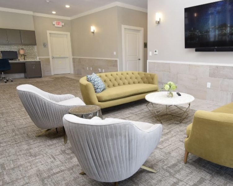 A modern lounge with a yellow sofa, gray armchairs, sleek flooring by a commercial flooring company, and a wall-mounted TV.