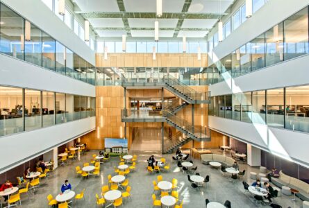 Spacious modern atrium with new flooring installation, large windows, central staircase, and people seated at yellow and white tables.