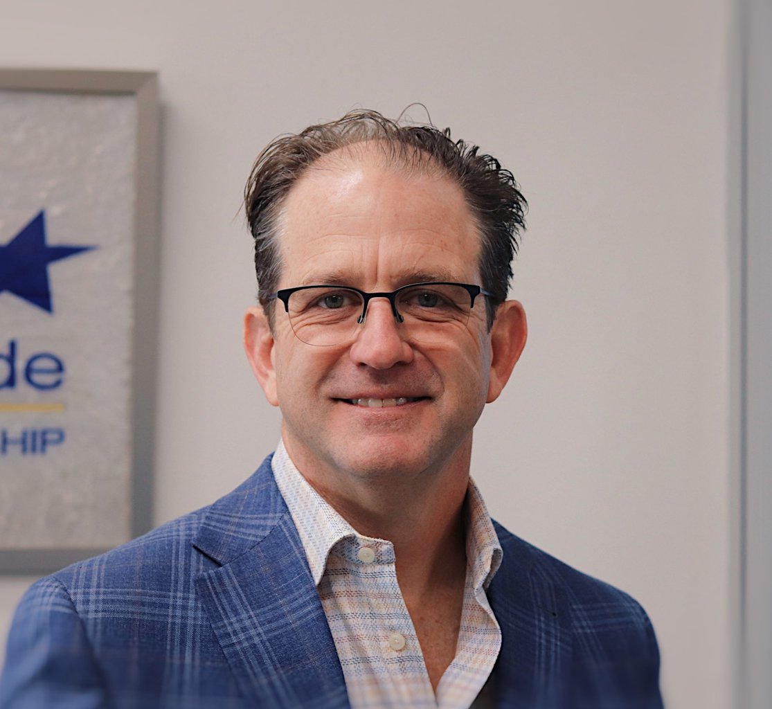 A man wearing glasses and a blue plaid blazer stands indoors at a commercial flooring company, smiling at the camera.